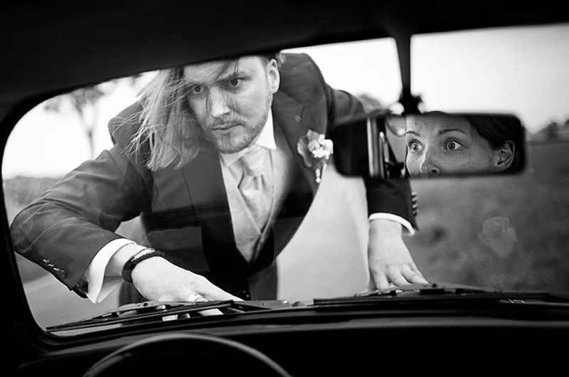 Hochzeitsbilder mal anders in Schwarz Weiß - der Bräutigam erschreckt die Braut, die im Auto sitzt.